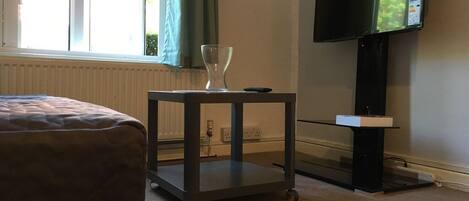 A hotel room with a bed, a small cart with a glass, a flat-screen TV mounted on the wall, and a window with curtains.