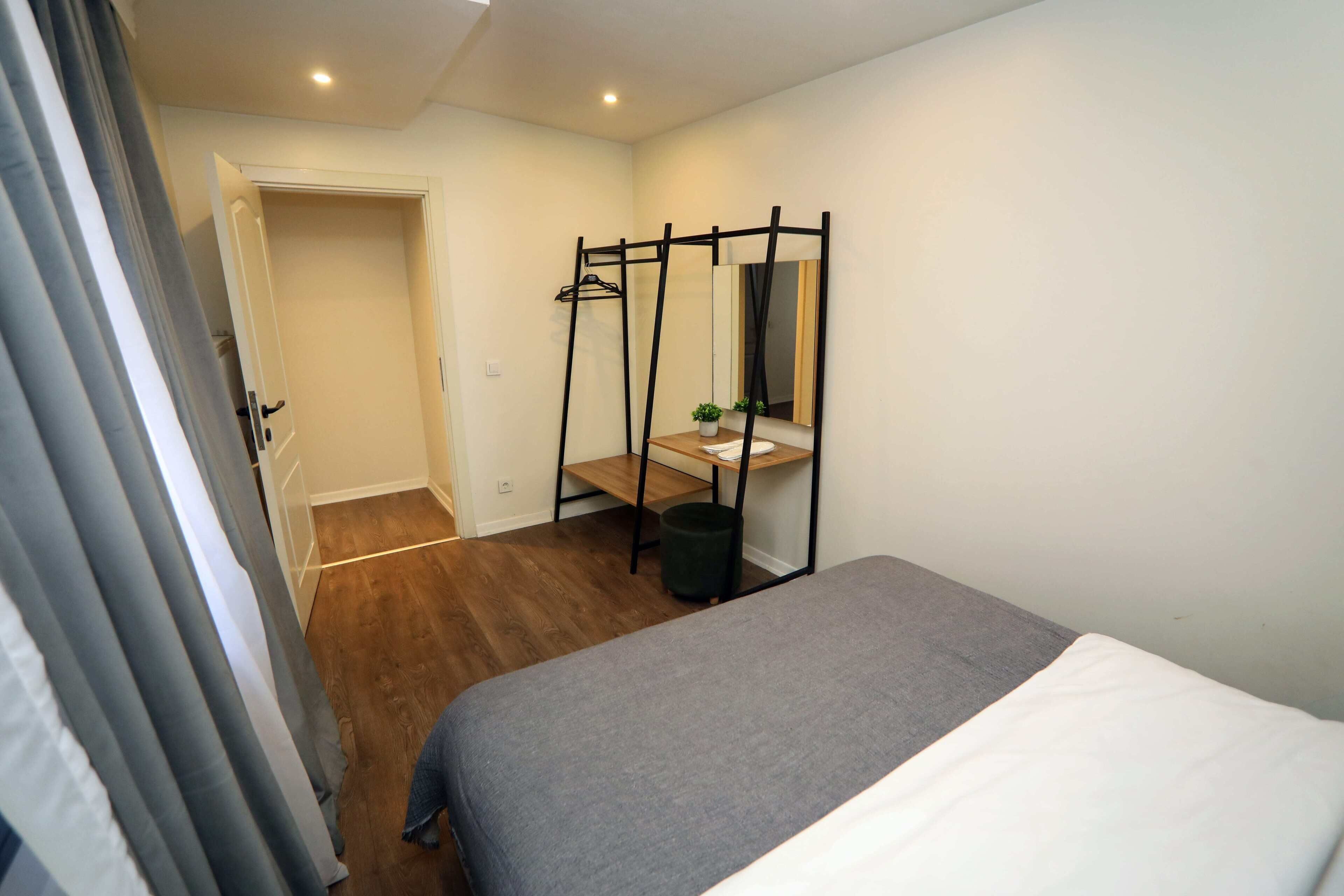 Une chambre à coucher comprenant un lit, une table de chevet, un miroir, un porte-vêtements et une porte donnant sur une autre pièce.