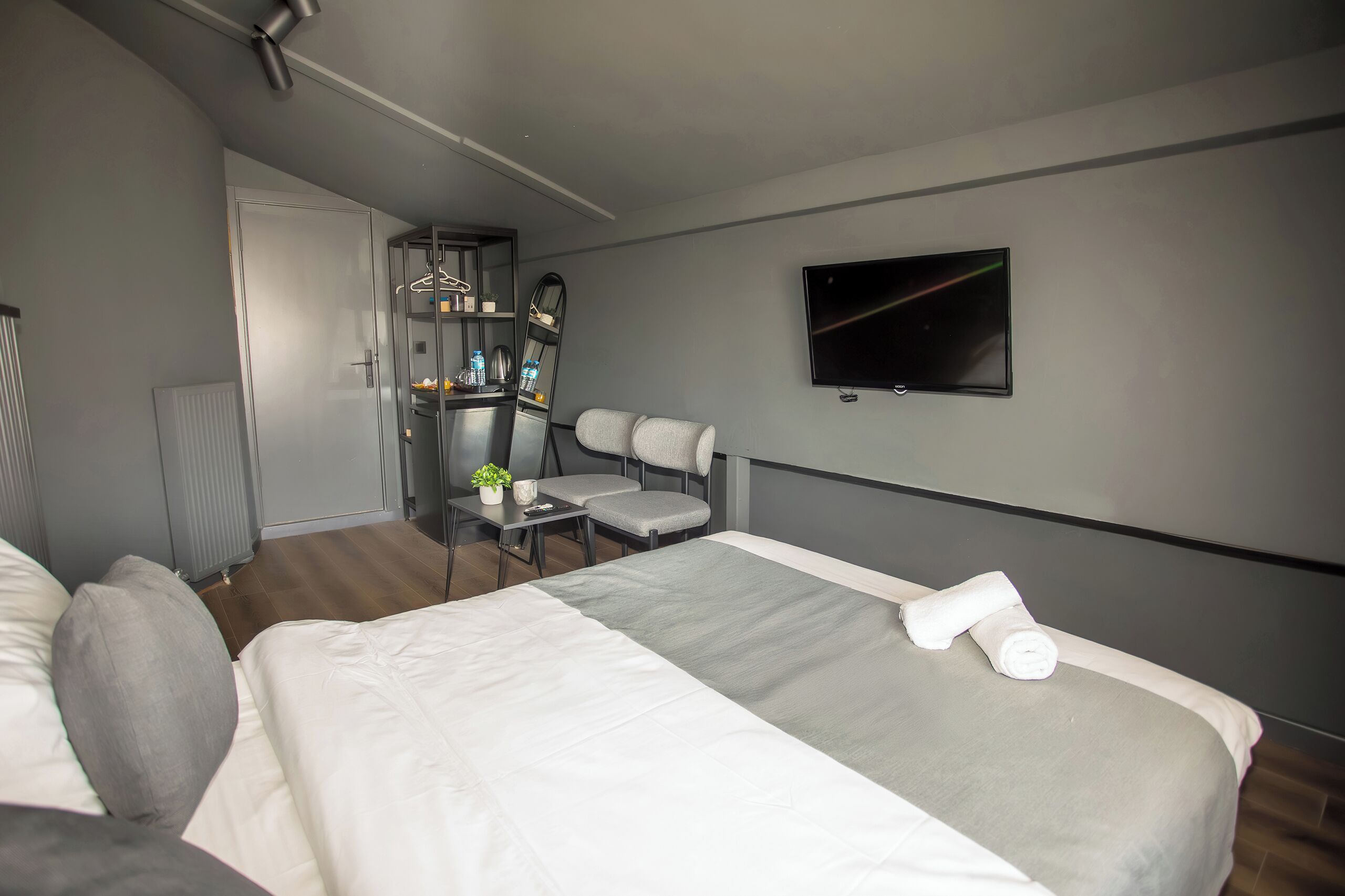 A modern hotel room with a large bed, a seating area with a small table and chairs, a TV mounted on the wall, and a wardrobe.