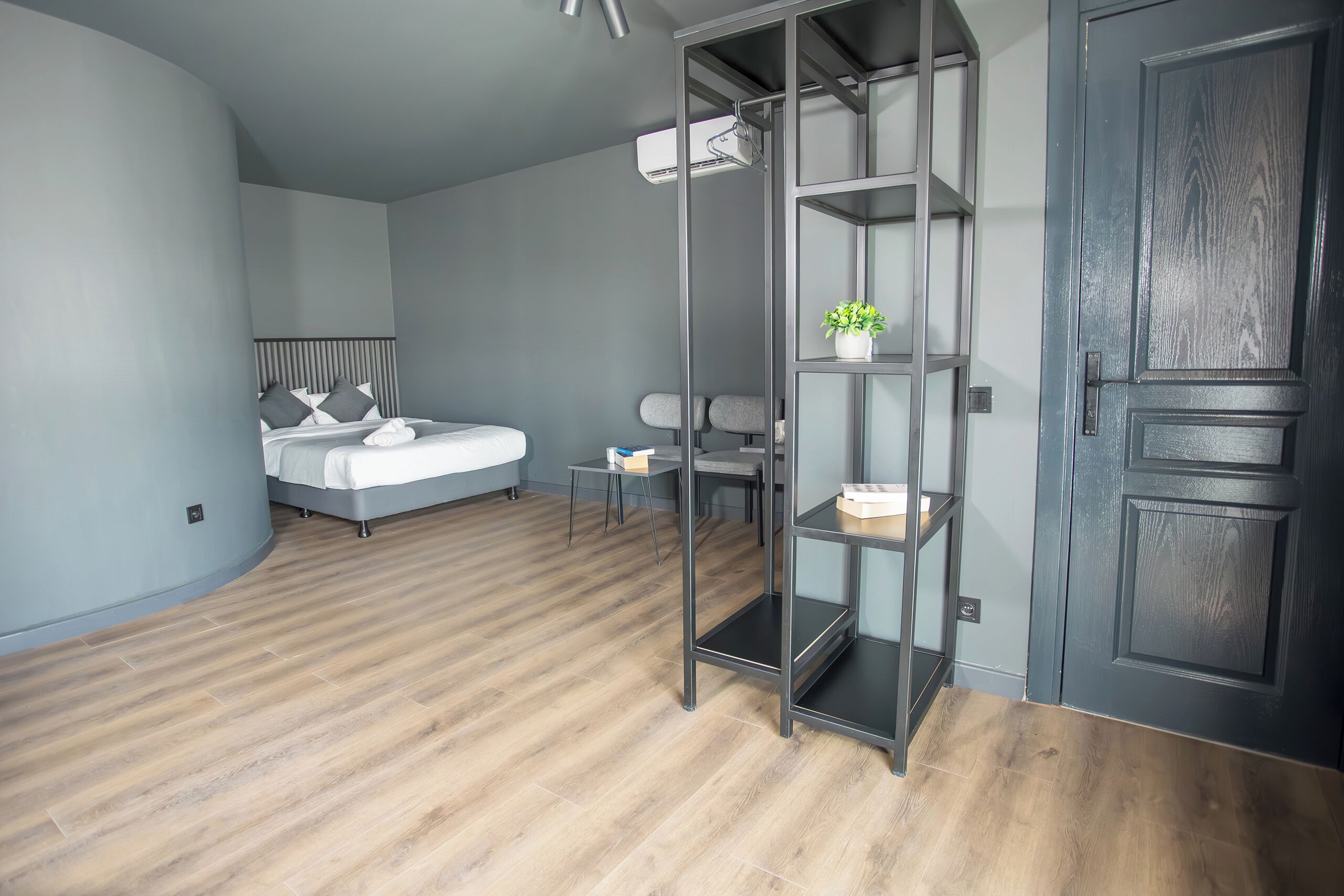 A modern hotel room with a bed, a desk, a chair, a wardrobe, and a shelf unit.