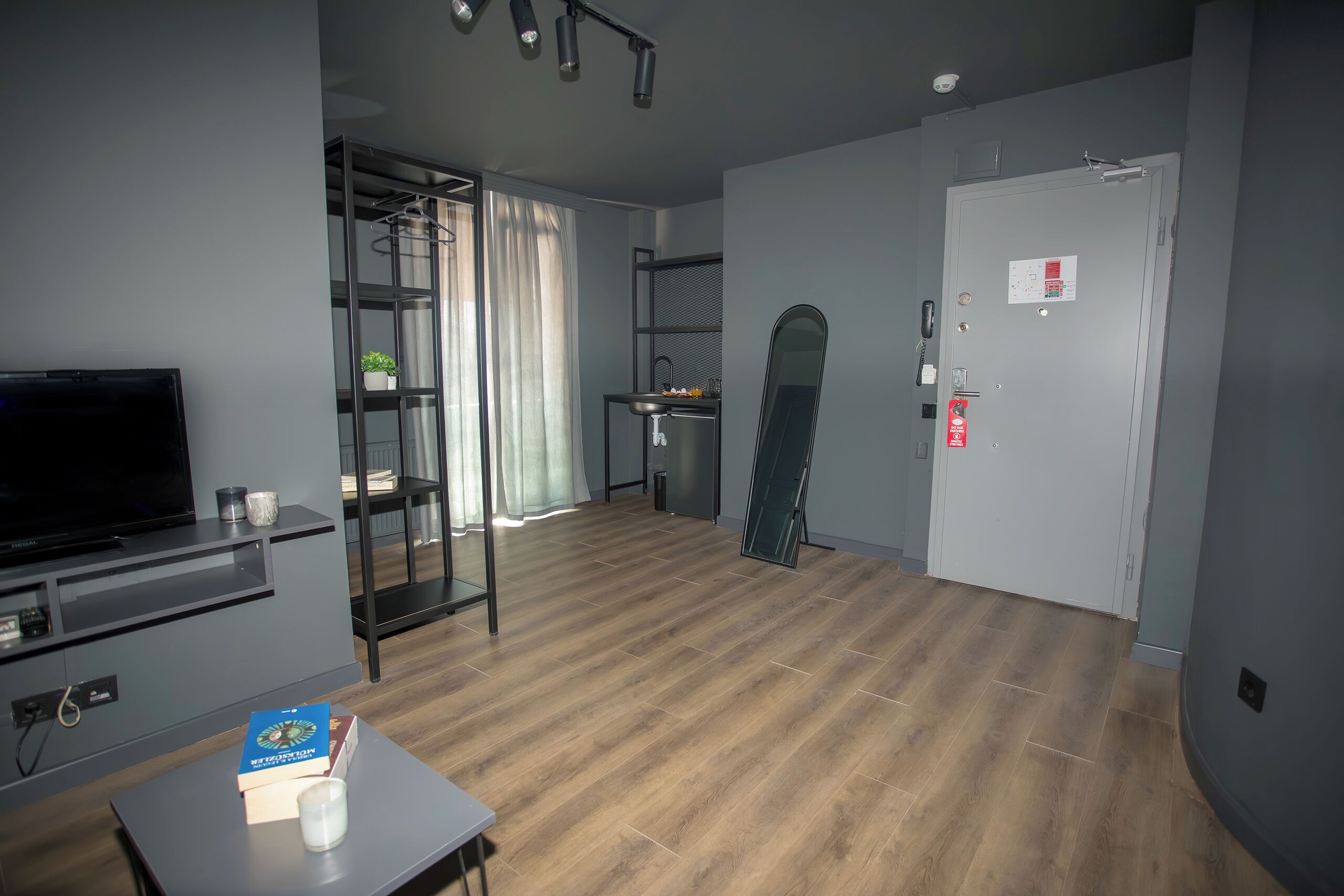 A modern living room with a flat-screen TV, a coffee table with a book and a glass, a wooden floor, a glass door leading to a balcony, and a white door with a fire safety sign.