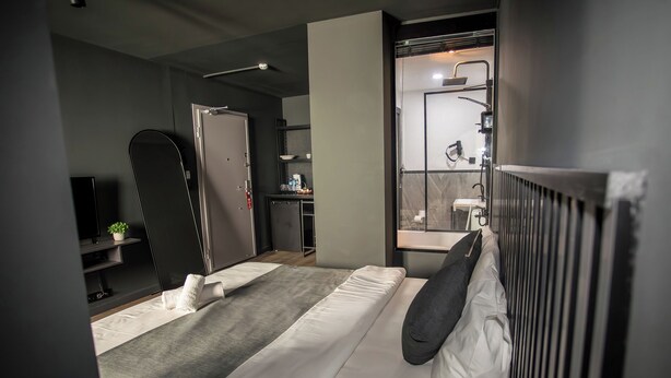 Una habitación de hotel moderna con una cama, un espejo grande, un baño con ducha y una pequeña cocina.
