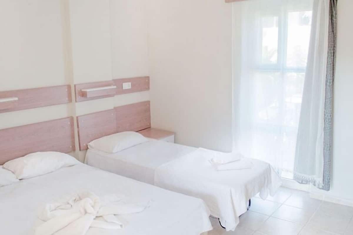 Una habitación con dos camas individuales, un cabecero de madera y un ventanal con cortinas traslúcidas.
