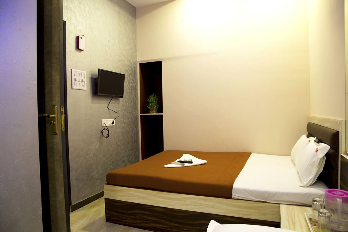A hotel room with a bed, a TV mounted on the wall, a power outlet, and a small shelf with a plant.
