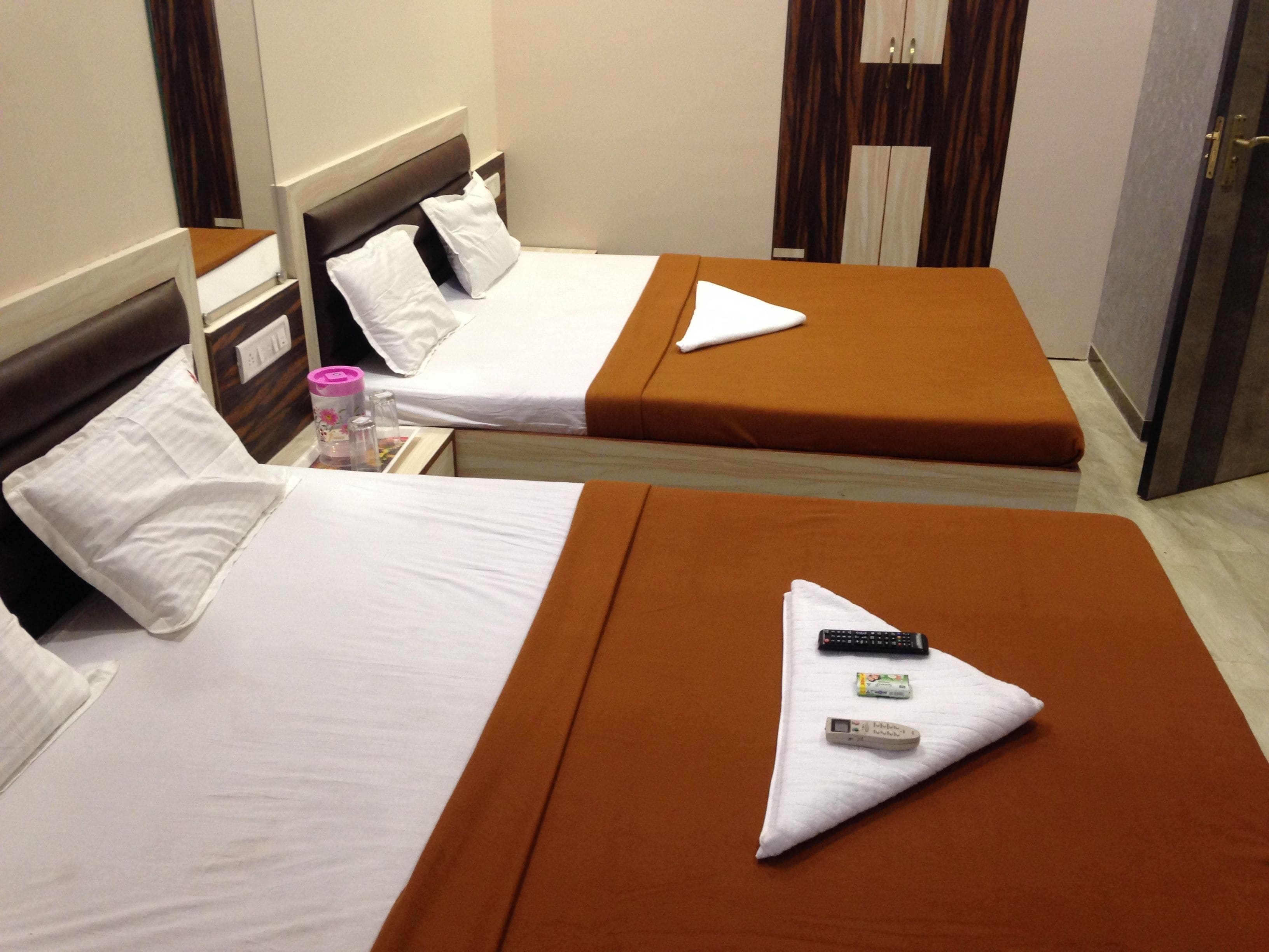 Two single beds with brown headboards and white bedding, a bedside table with a pink container, and a door with a mirror.