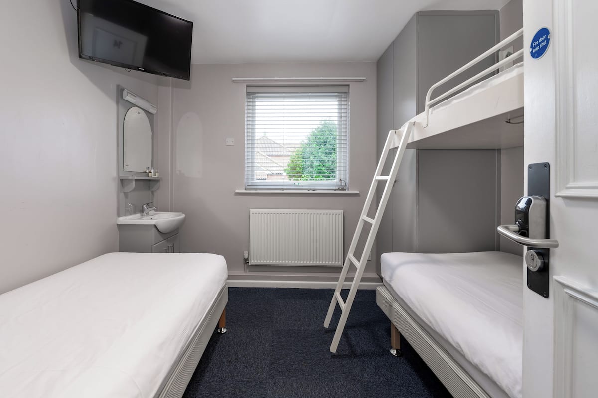 Una habitación pequeña y limpia con dos camas individuales, televisión integrada, ventana con persianas, radiador y una puerta blanca con un letrero azul.