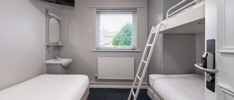 Una habitación pequeña y limpia con dos camas individuales, televisión integrada, ventana con persianas, radiador y una puerta blanca con un letrero azul.