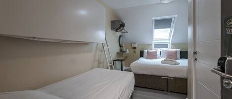 A compact hotel room with two single beds, a small bedside table, a mirror, a window, and a door.