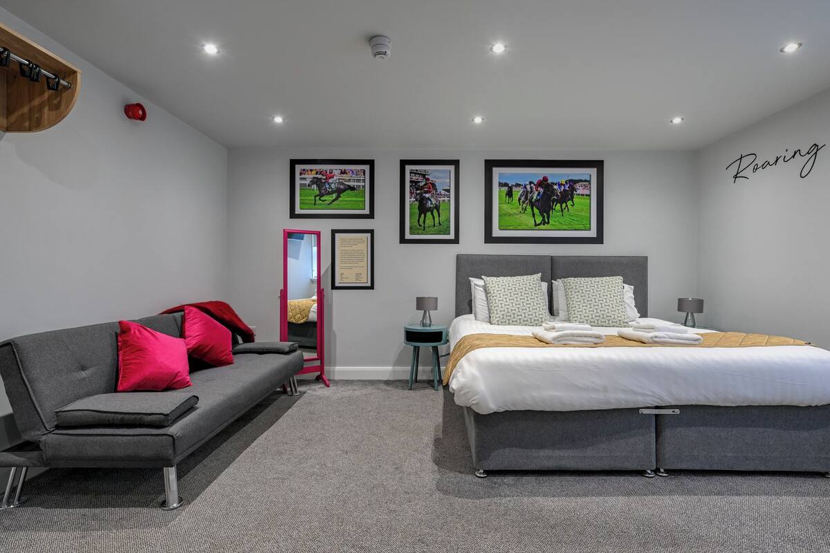 Una habitación de hotel moderna con un sofá gris, una cama grande y tres carteles enmarcados de carreras de caballos en la pared.