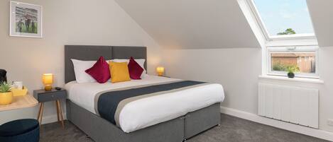 A modern bedroom with a grey bed, red and yellow pillows, a bedside table with a lamp, a small desk with a plant, and a view through a skylight.
