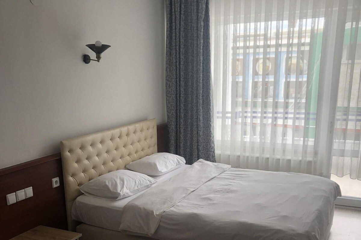 A hotel room with a large bed, a bedside table, a wall-mounted lamp, and a window with curtains.
