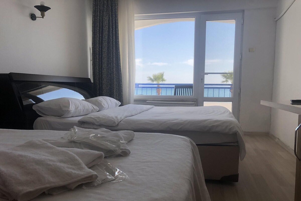 Une chambre d’hôtel avec deux lits, une fenêtre donnant sur la mer, un ventilateur de plafond et une lampe fixée au mur.