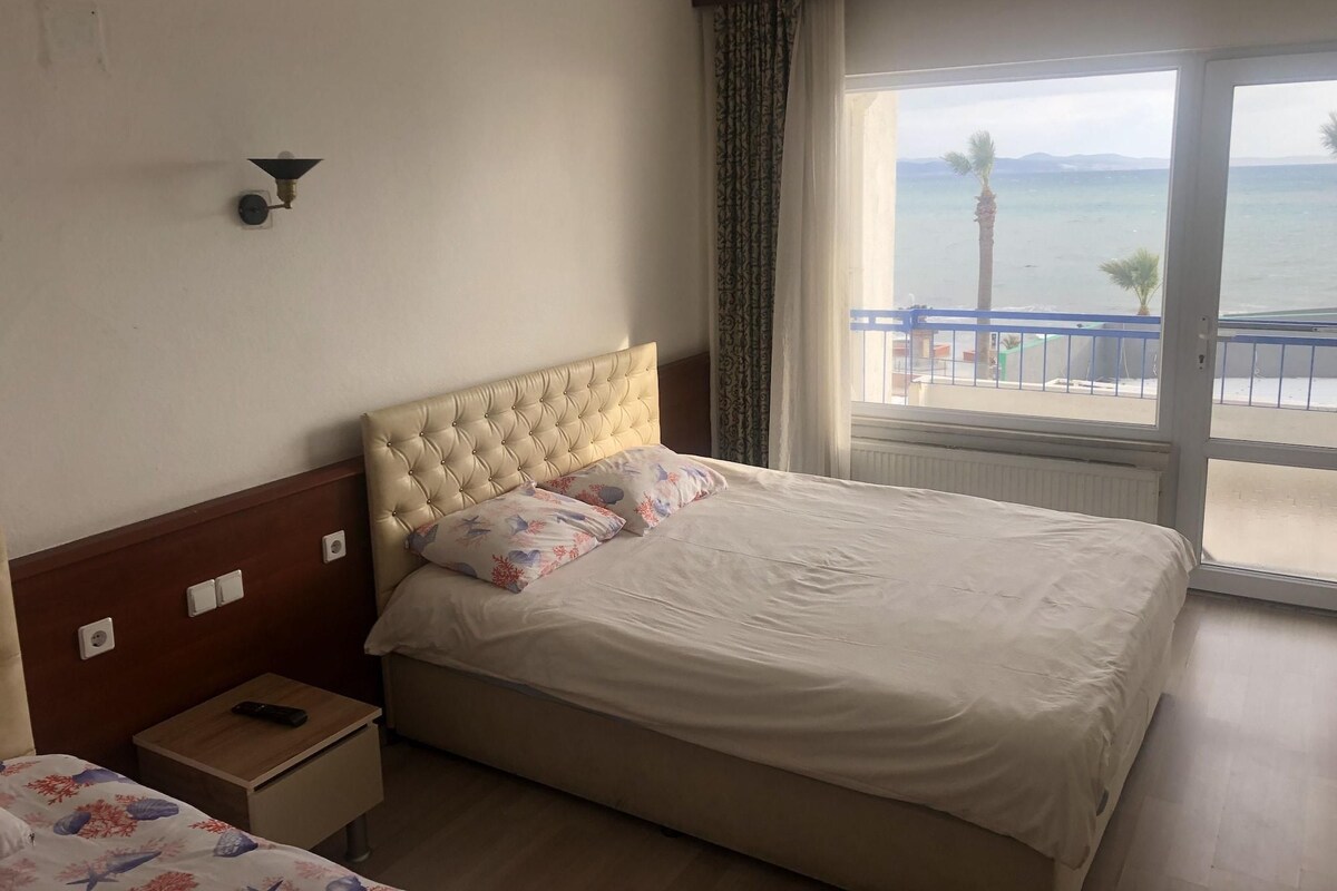 A hotel room with a bed, a bedside table with a remote control, a window with a view of the sea, and a wall-mounted light fixture.