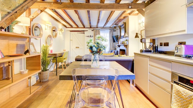 A modern kitchen with wooden beams, a dining area with clear chairs, and a central island with a vase of flowers. - Cartref (Farnham)
