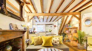 A cozy room with exposed wooden beams, a sofa with yellow cushions, a round mirror, and a bookshelf. - Cartref (Farnham)