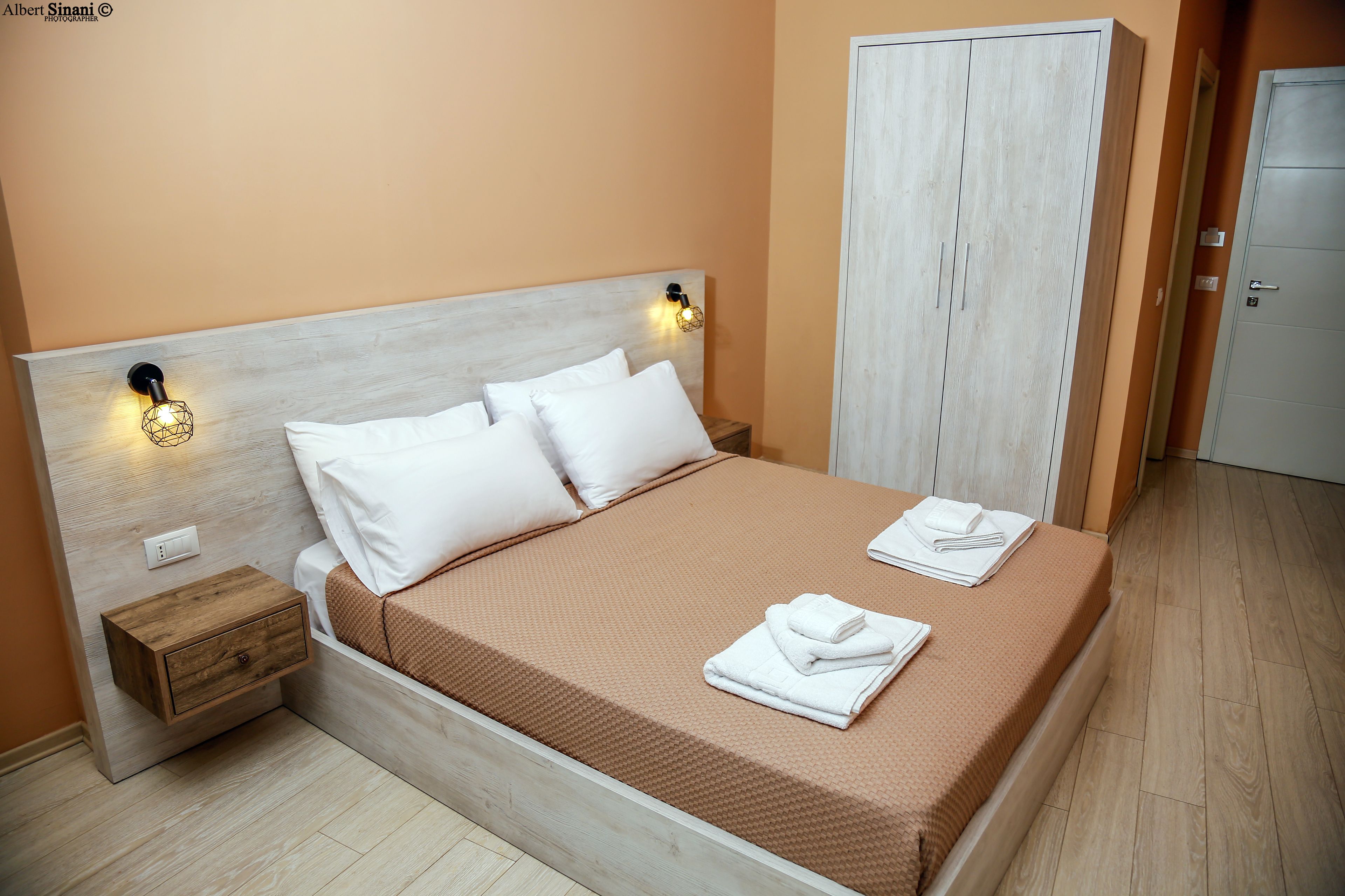 A neatly made bed with white linens and a brown bedspread, a wooden nightstand with a drawer, and a wardrobe with a light wood finish.