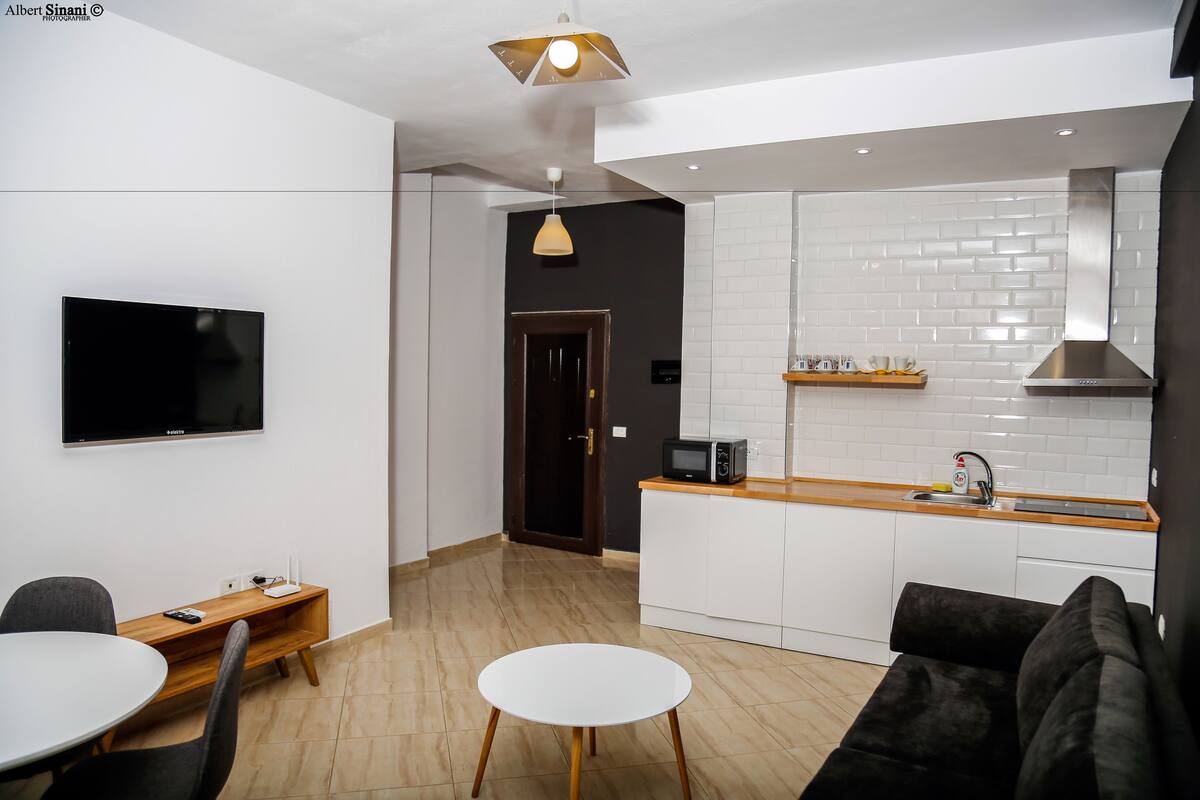 A modern kitchen with white cabinets, a wooden countertop, and a built-in microwave. There is a dining area with a round table and chairs, and a black sofa.