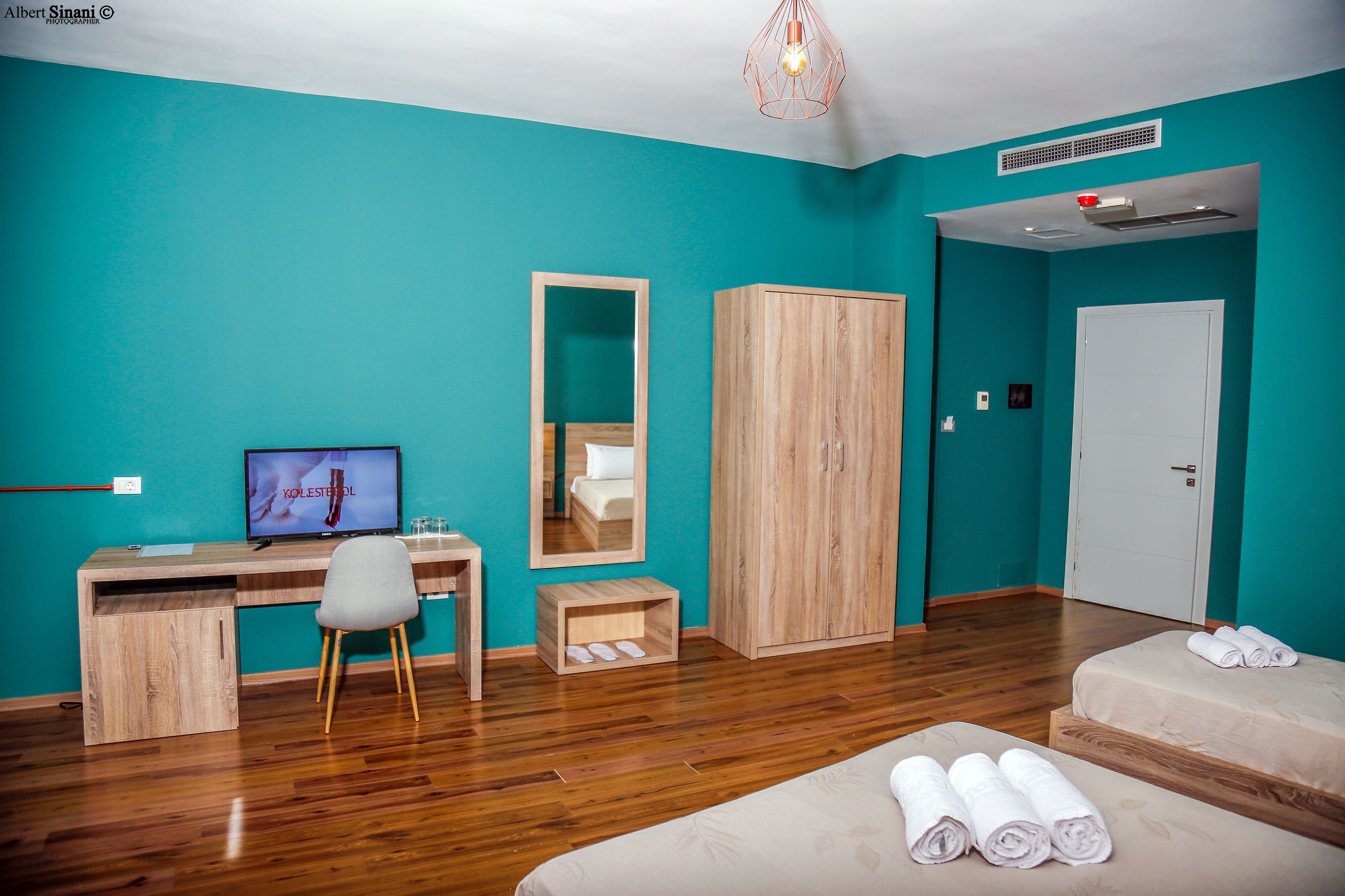 Una habitación de hotel moderna con paredes color turquesa, un escritorio de madera, un televisor, un espejo, un armario, una cama con toallas y suelo de madera.