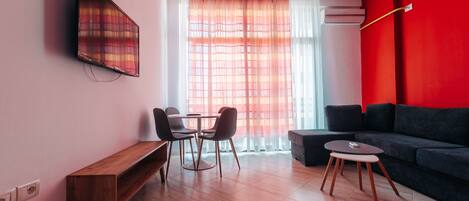 Una sala de estar moderna con un televisor de pantalla plana montado en la pared, una zona de comedor con una mesa y sillas de madera, un sofá y una pequeña mesa redonda.