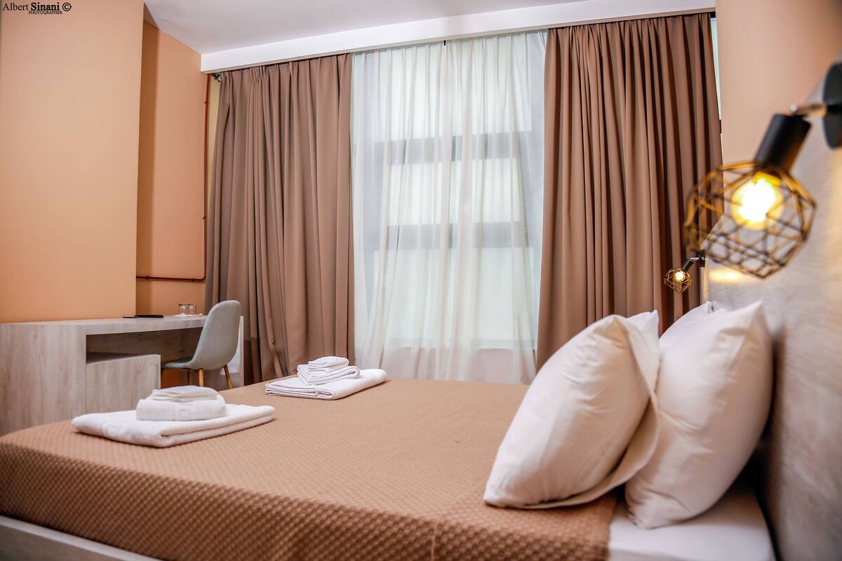 A hotel room with a bed, a desk, a chair, and a large window with curtains.