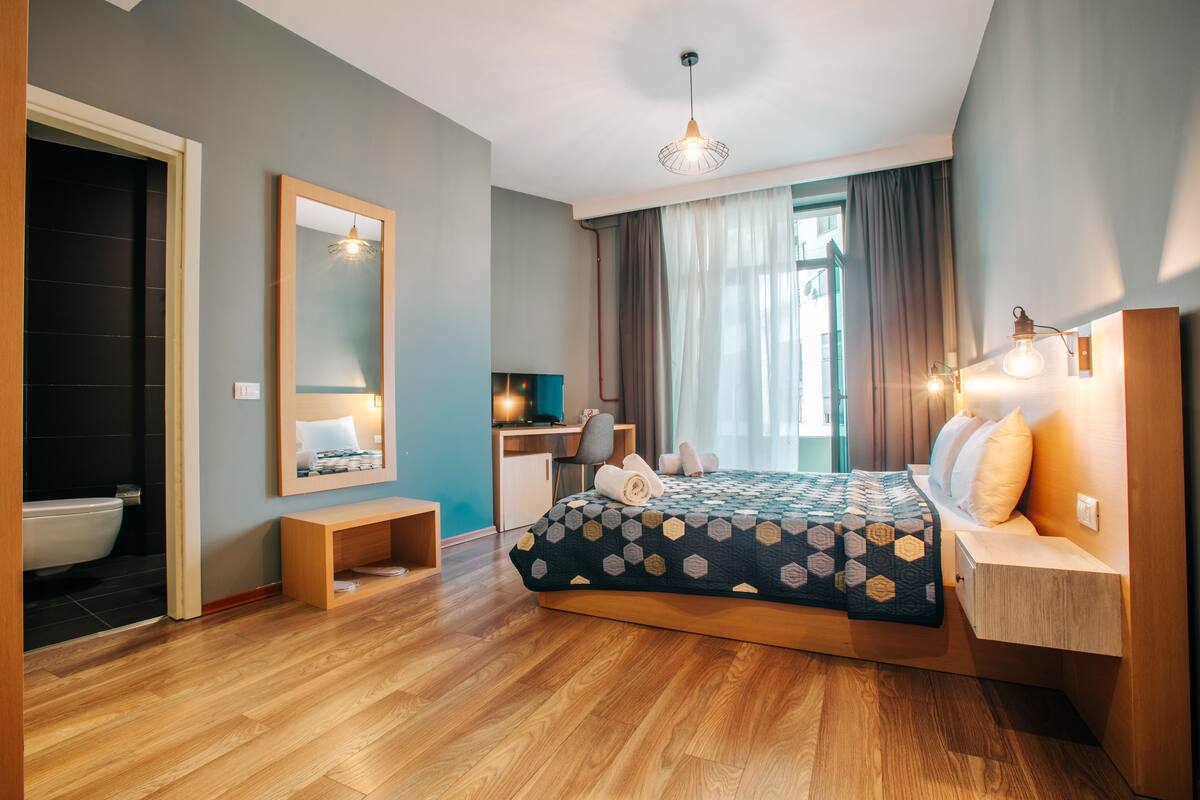 A modern bedroom with a wooden floor, a bed with a geometric-patterned duvet, a bedside table, a desk with a chair, a mirror, and a window with curtains.