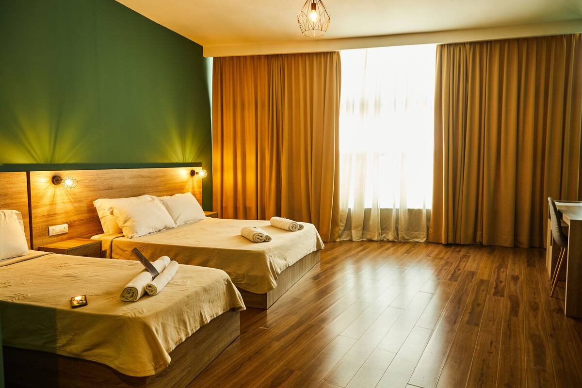 A hotel room with two beds, wooden flooring, a large window with curtains, and a chandelier.