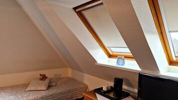 A room with a sloped ceiling, a bed with a floral bedspread, a bedside table with a coffee maker, and a television.