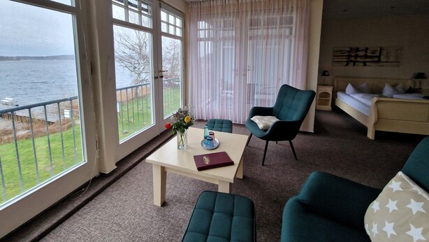A room with a view of the sea, a table with a vase and book, a green armchair, and a bed with pillows. - Faehrhaus Niederkleveez (Bösdorf)