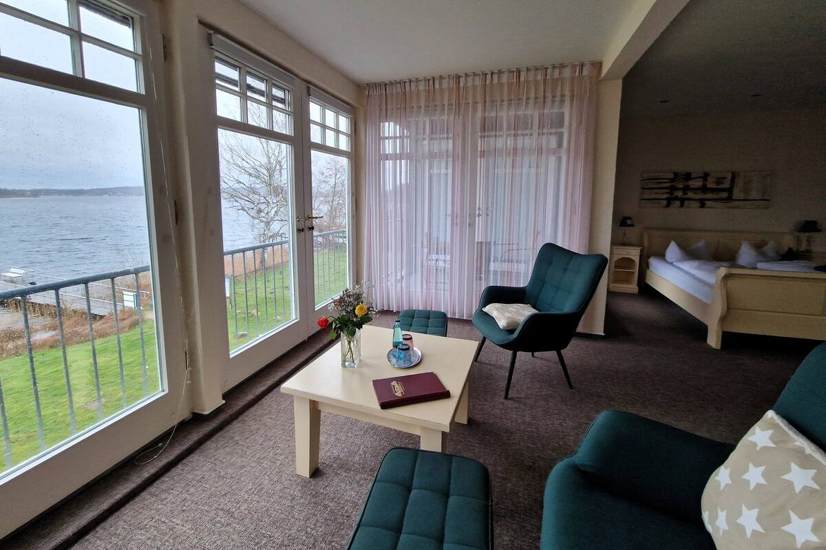 A room with a view of the sea, a table with a vase and book, a green armchair, and a bed with pillows.
