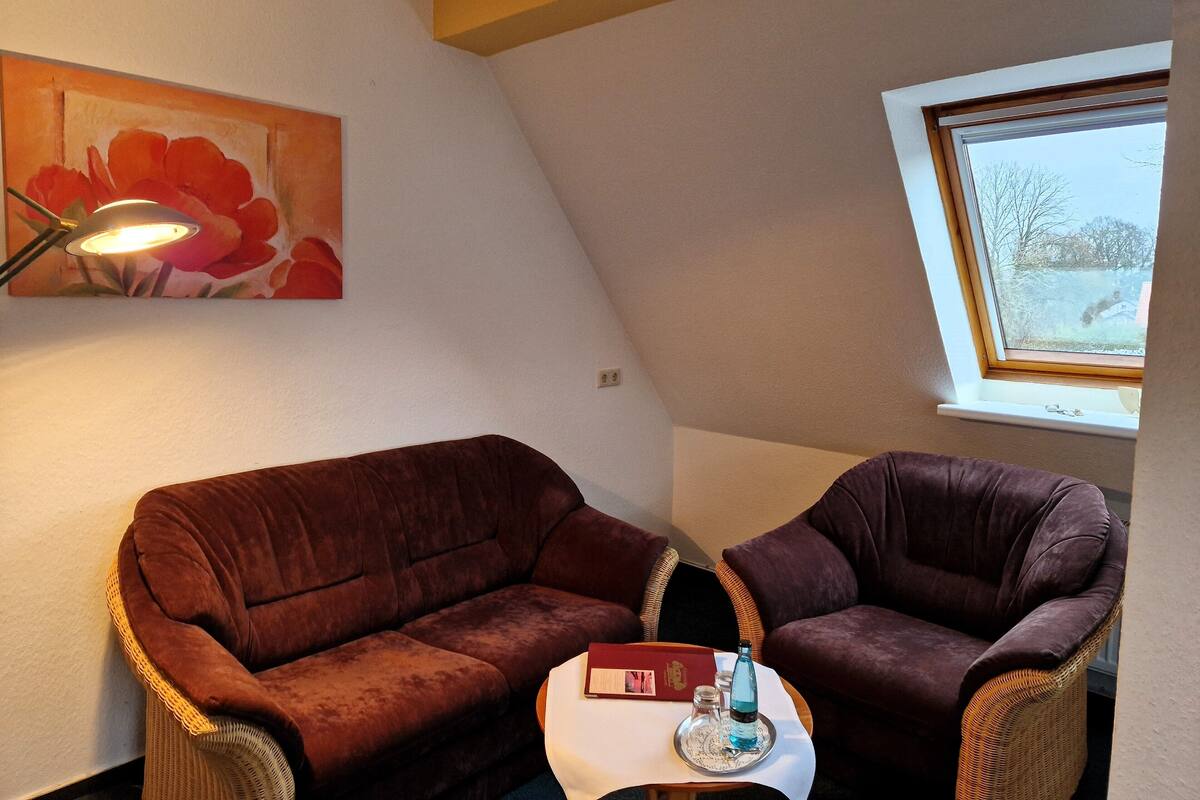 A cozy room with a brown sofa and a wicker chair, a small table with a book and a bottle, a painting on the wall, and a window with a view of trees.