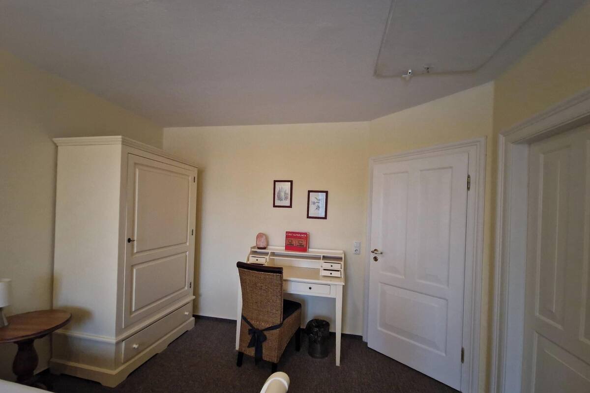 A hotel room with a large wardrobe, a desk with a chair, and two framed pictures on the wall.