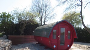 A red, cylindrical structure resembling a house with a door and windows, situated on a sandy area with a stone wall and trees in the background. - Faehrhaus Niederkleveez (Bösdorf)