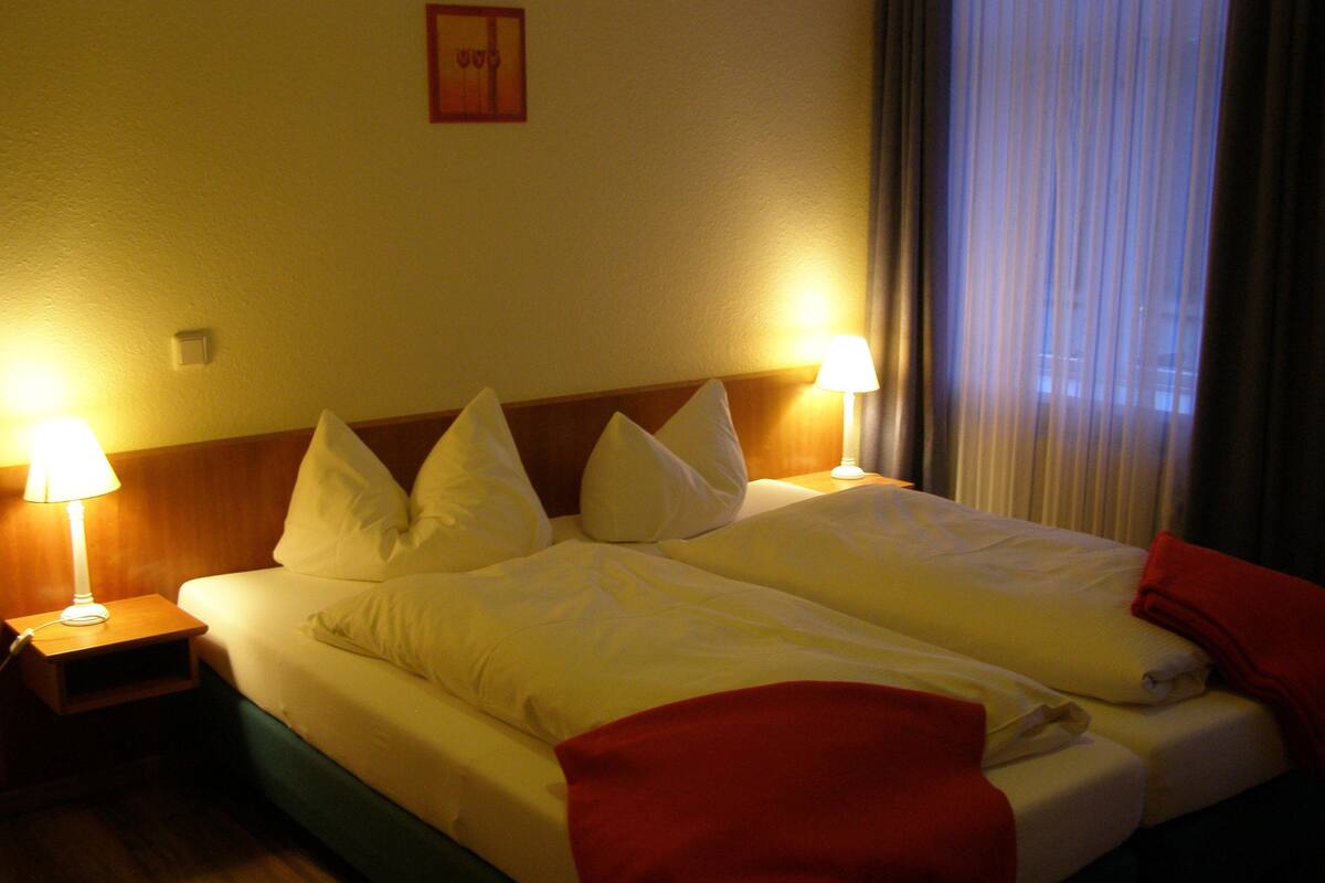 A hotel room with a bed, two bedside lamps, a nightstand, and a window with curtains.