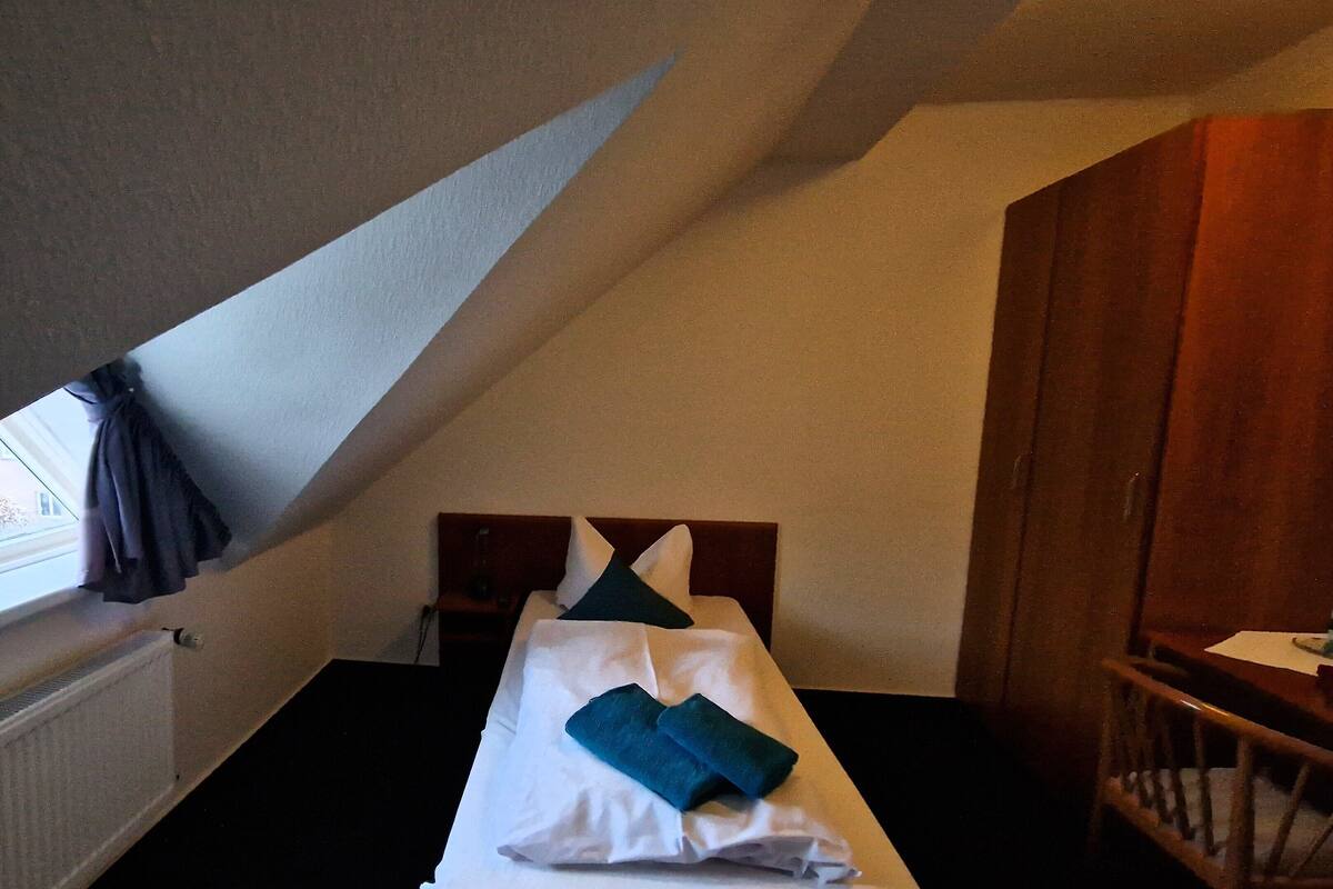 A small, dimly lit room with a single bed, a wooden chair, and a window with curtains.