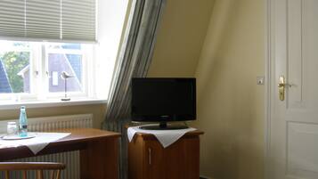 Una habitación con un escritorio de madera, un televisor sobre un mueble, una ventana con persianas y una puerta.