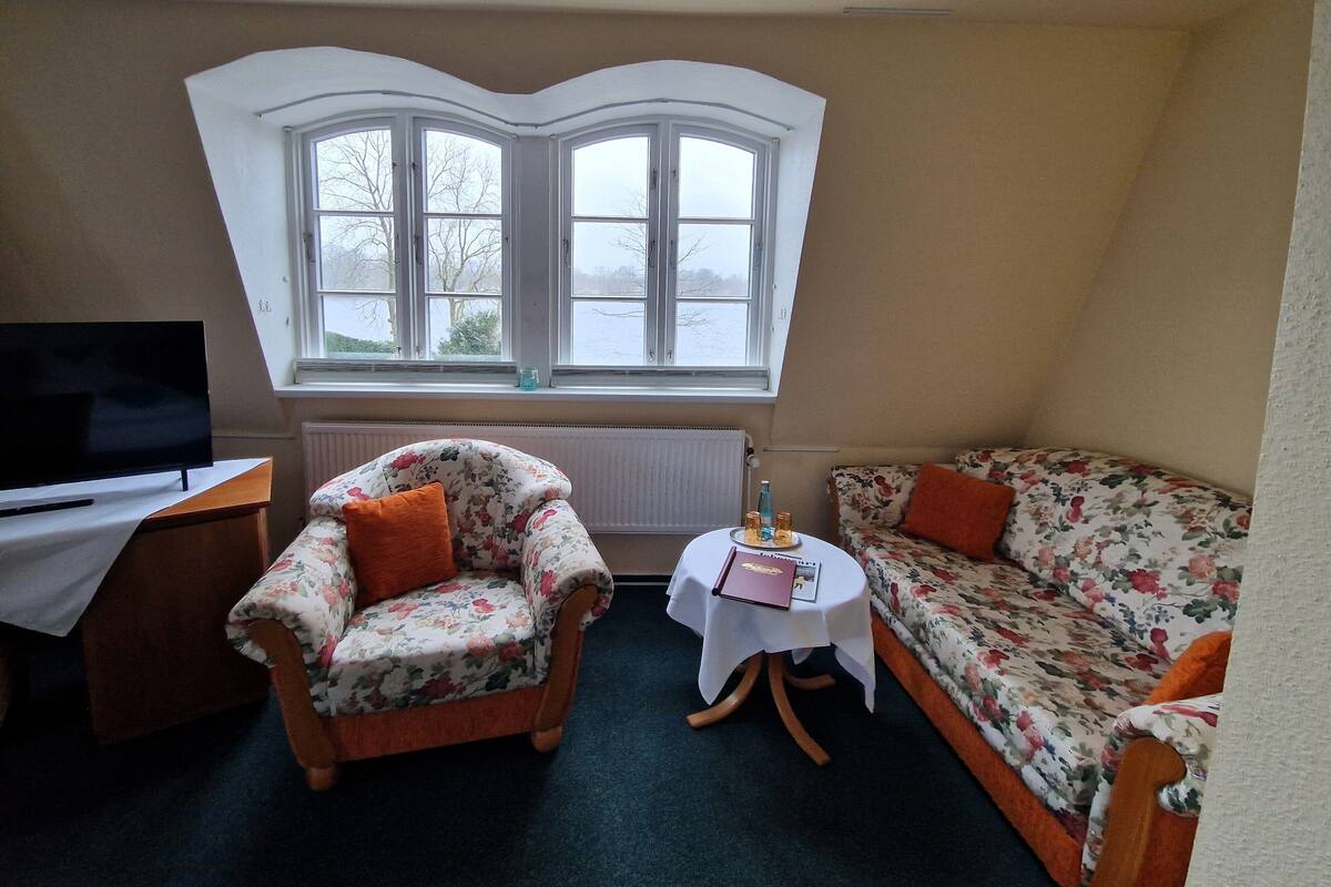 A cozy room with a large bay window, a television on a wooden table, a floral armchair, a sofa, and a small round table with a book and a bottle.