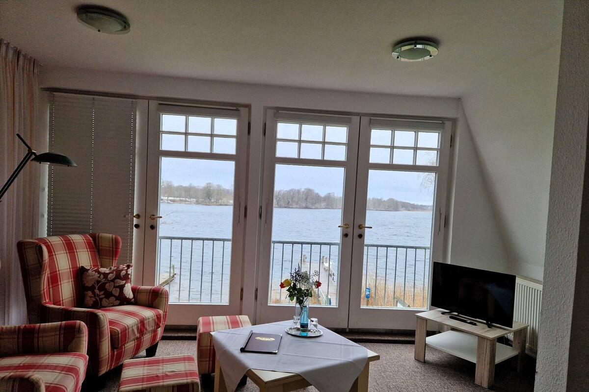A room with a view of a lake, featuring a plaid sofa, a small table with a vase, and a TV on a stand.