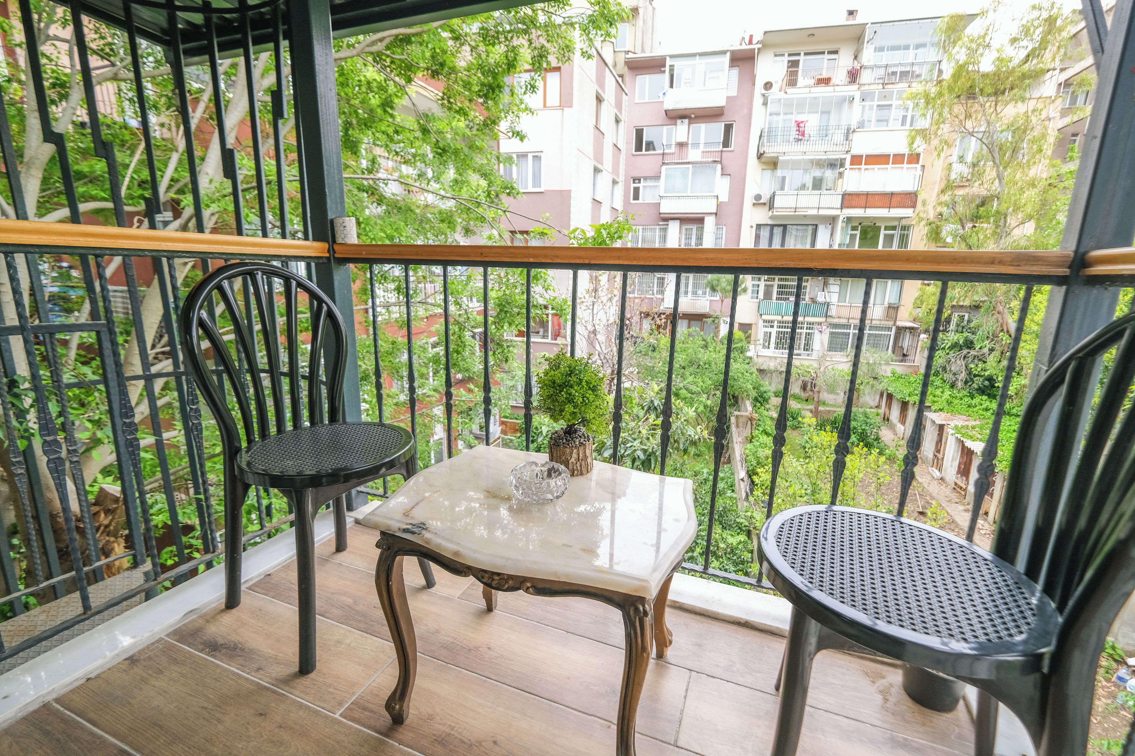 A balcony with a table and two chairs, a small plant, and a view of a residential area with multi-story buildings.