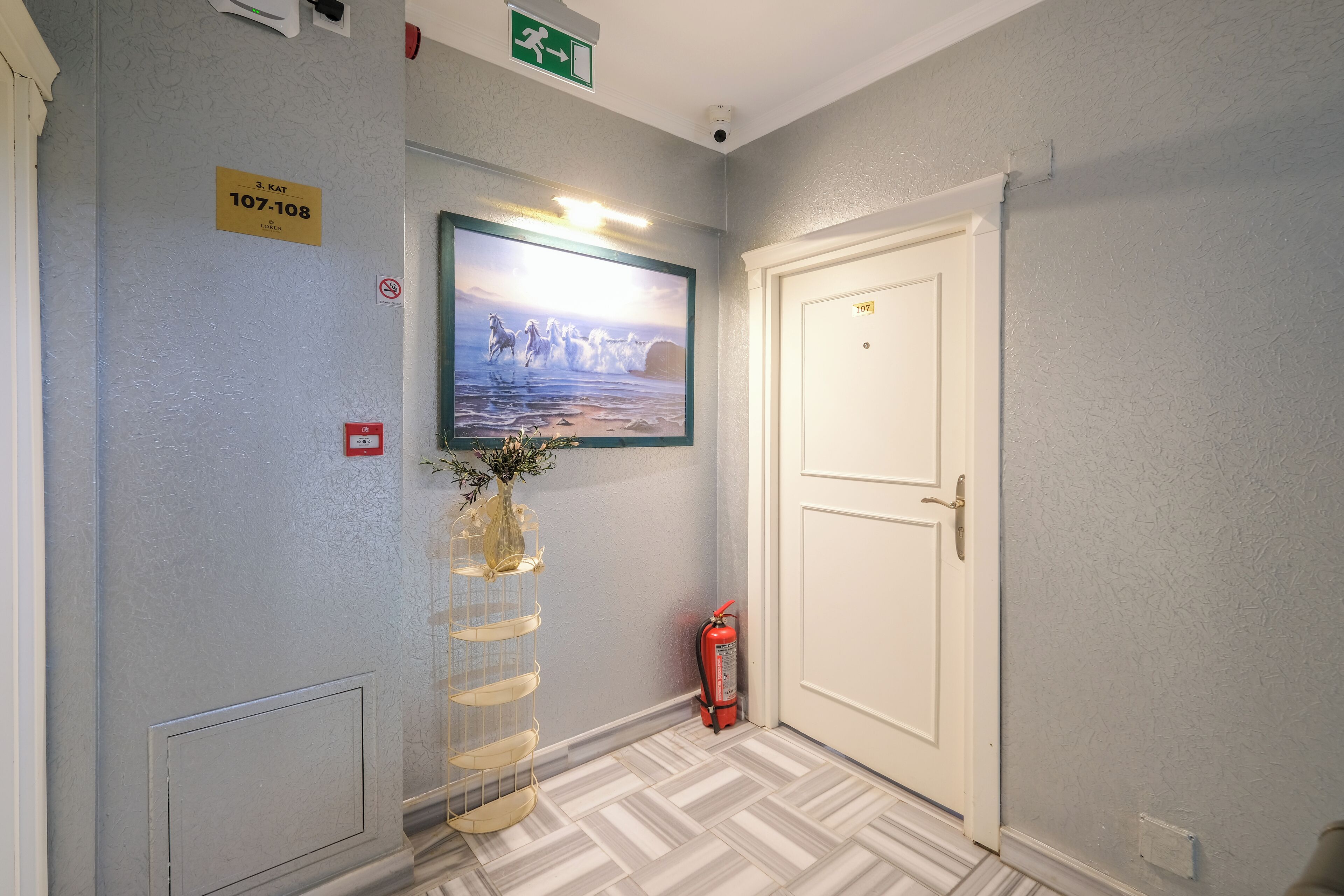 A hallway with a framed picture, a fire extinguisher, and a door marked 107-108.