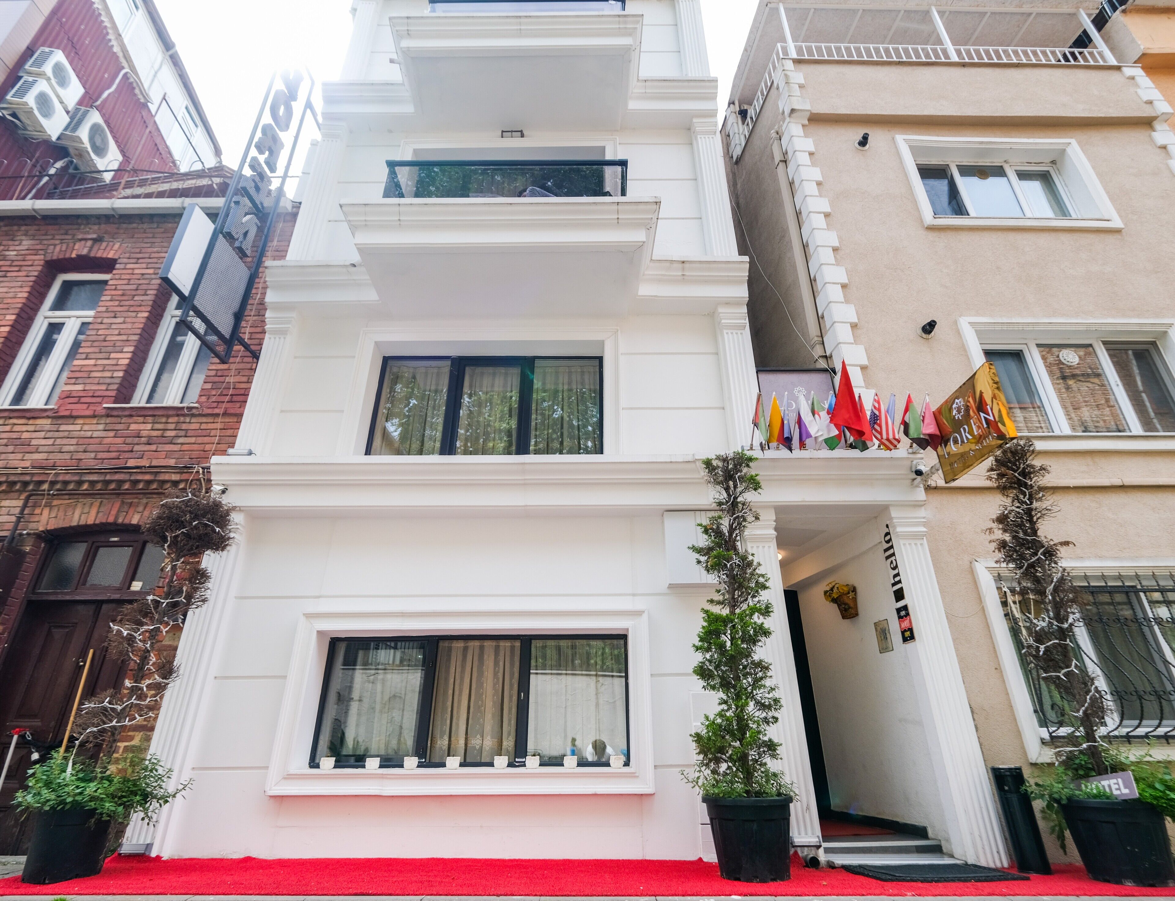 Un hotel de varios pisos con fachada blanca, balcones adornados con banderas y una entrada con alfombra roja.