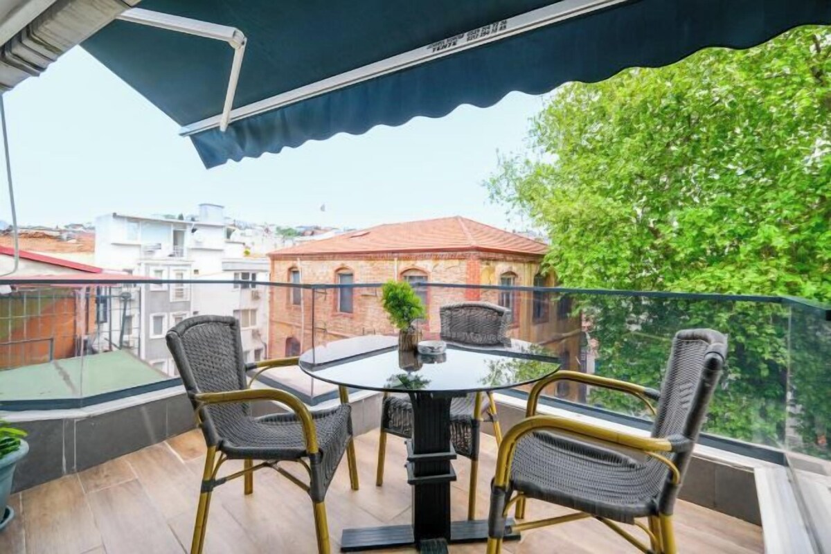 A balcony with a glass table and chairs, a potted plant, and a view of buildings and trees.