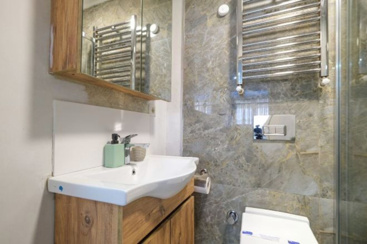 A bathroom with a marble wall, a wooden vanity, a white sink, a mirror, and a glass shower enclosure.