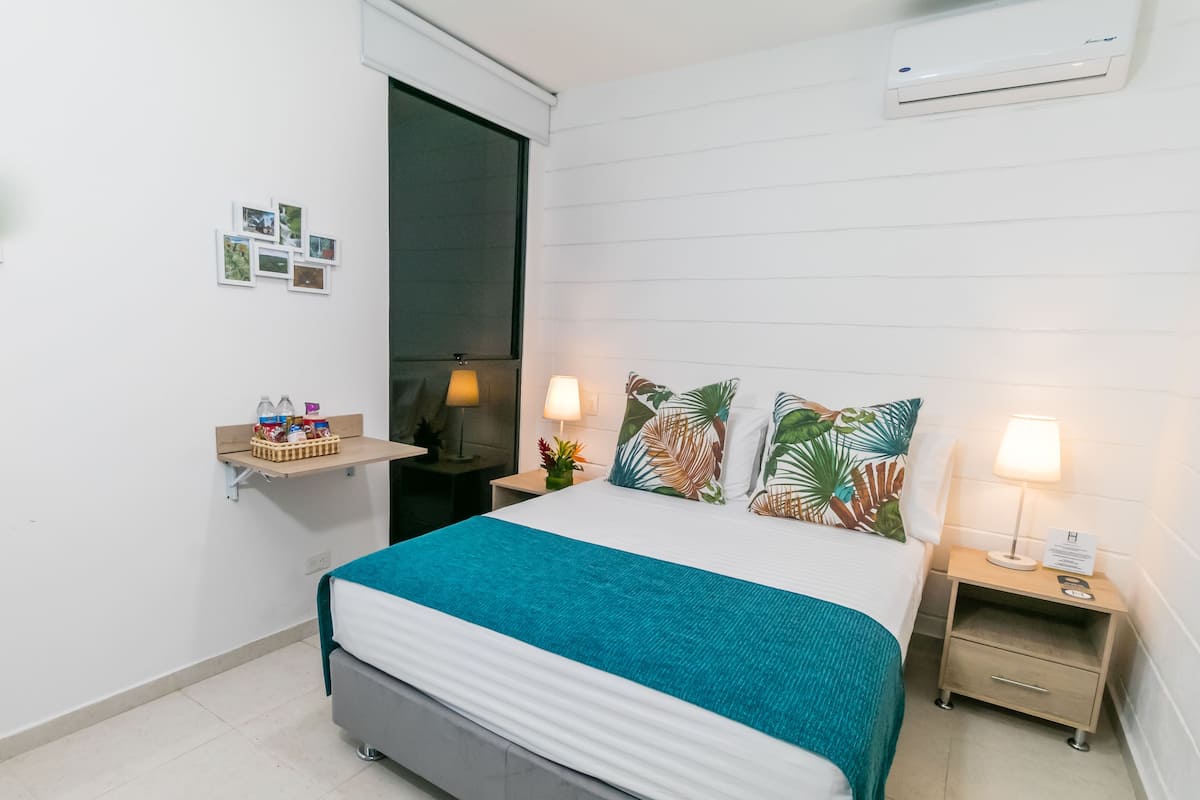 Habitación de hotel ordenada con cama, mesitas de noche, un estante con artículos de aseo y aire acondicionado de pared.
