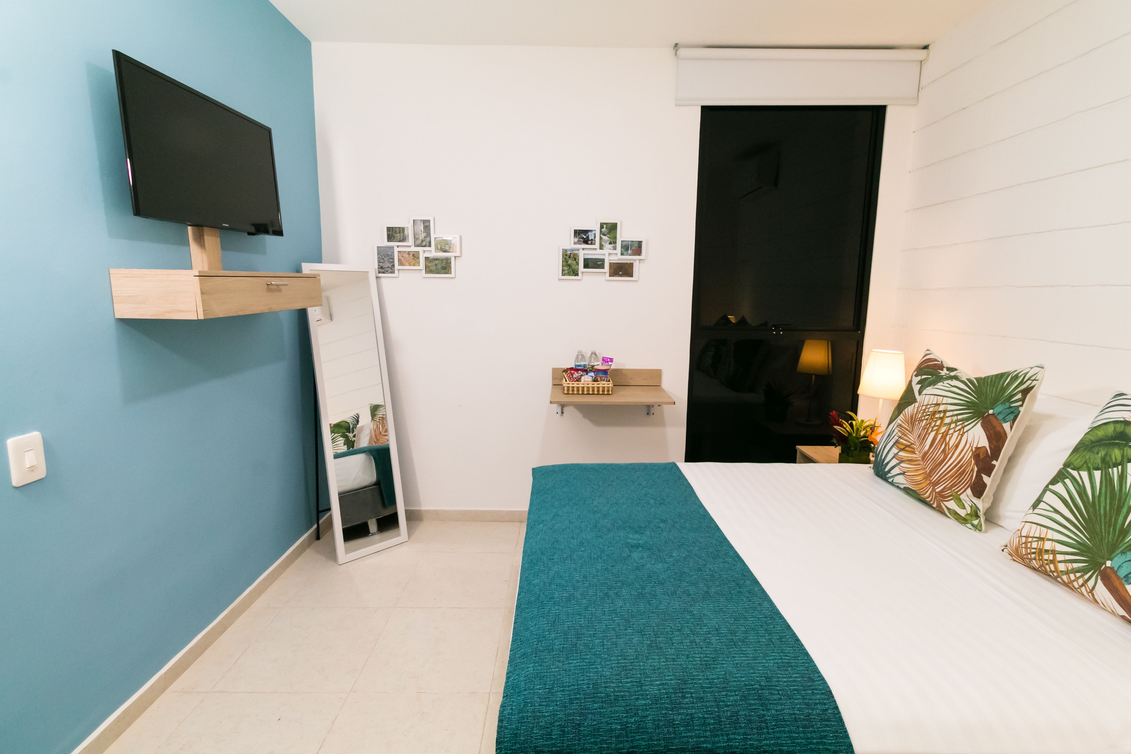 A bedroom with a bed, a TV mounted on the wall, a mirror, a shelf with photos, and a door leading to another room.