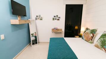 A bedroom with a bed, a TV mounted on the wall, a mirror, a shelf with photos, and a door leading to another room.