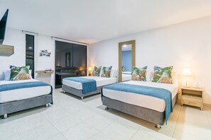Habitación de hotel con tres camas, cada una con una manta azul y almohadas tropicales. Hay una mesita de noche de madera con una lámpara y un espejo en la pared.