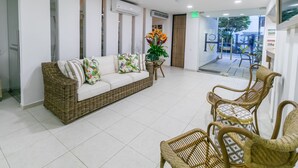 Un área de recepción con un sofá y sillones de mimbre, una mesita con un jarrón de flores y un ventanal que da al exterior.