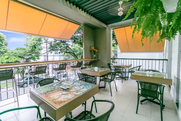Un café al aire libre con mesas y sillas, una planta colgante y vistas a la calle y árboles.