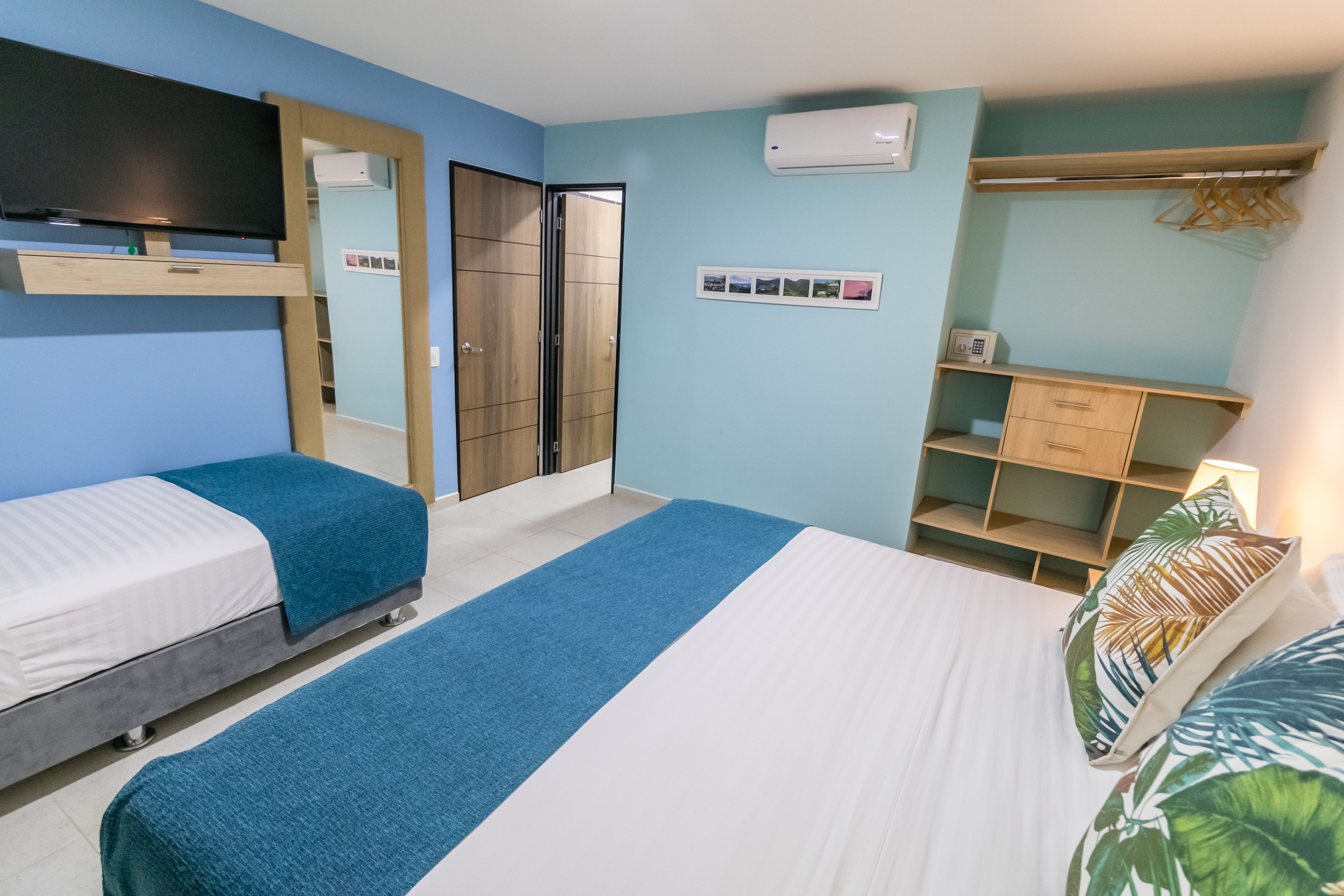 Habitación de hotel con dos camas individuales, televisión, armario, espejo, estante con lámpara y aire acondicionado instalado en la pared.