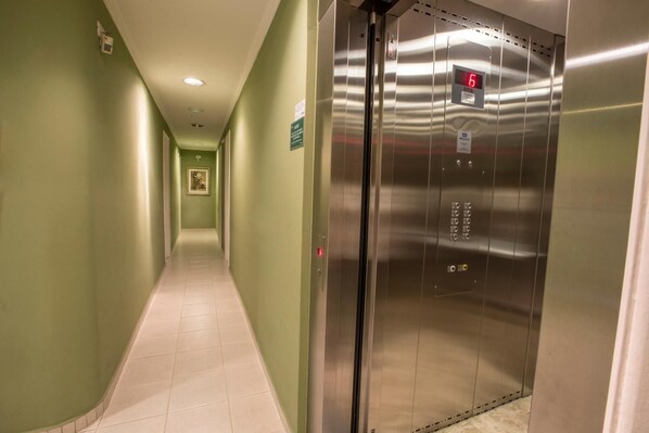 Un pasillo con una pared verde y un ascensor metálico que muestra el número 6 en su pantalla digital.